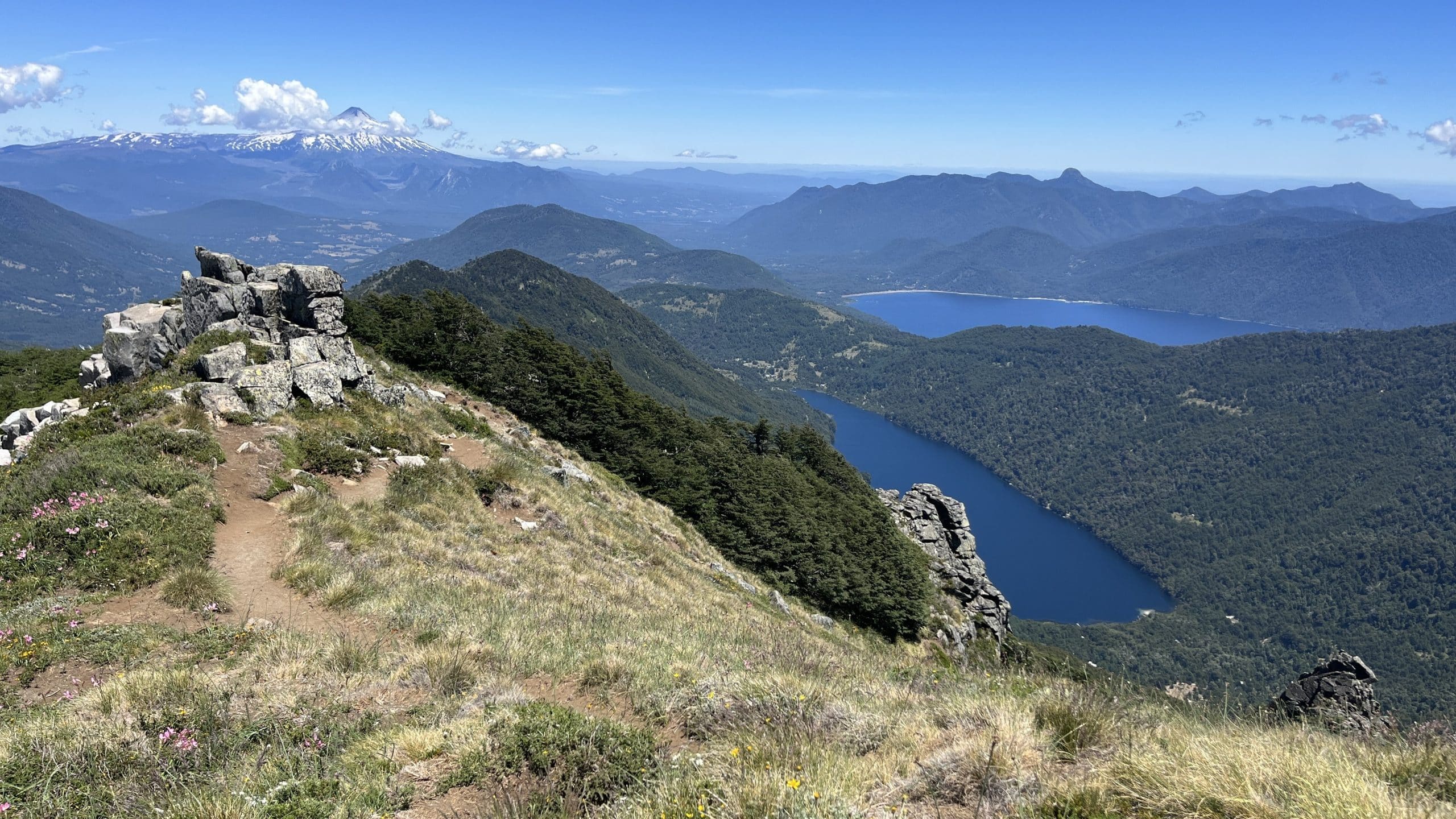A panoramic view from a rocky mountain ridge in Huerquehue National Park, overlooking deep blue lakes and densely forested valleys with a snow-capped volcano visible on the distant horizon.