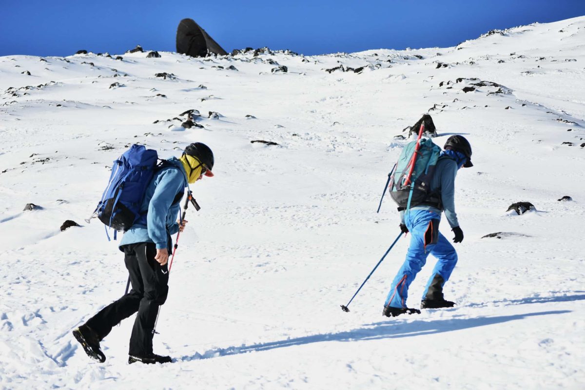 Two mountaineers equipped with backpacks and trekking poles ascending a steep, snowy slope on the Villarrica volcano under a bright blue sky.